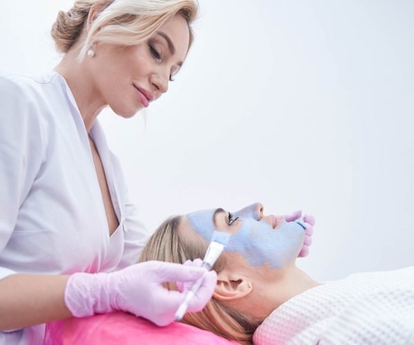 Focused beautician applying mask to face of calm young woman Cosmetician spreading blue clay paste over facial skin of client using two brushes